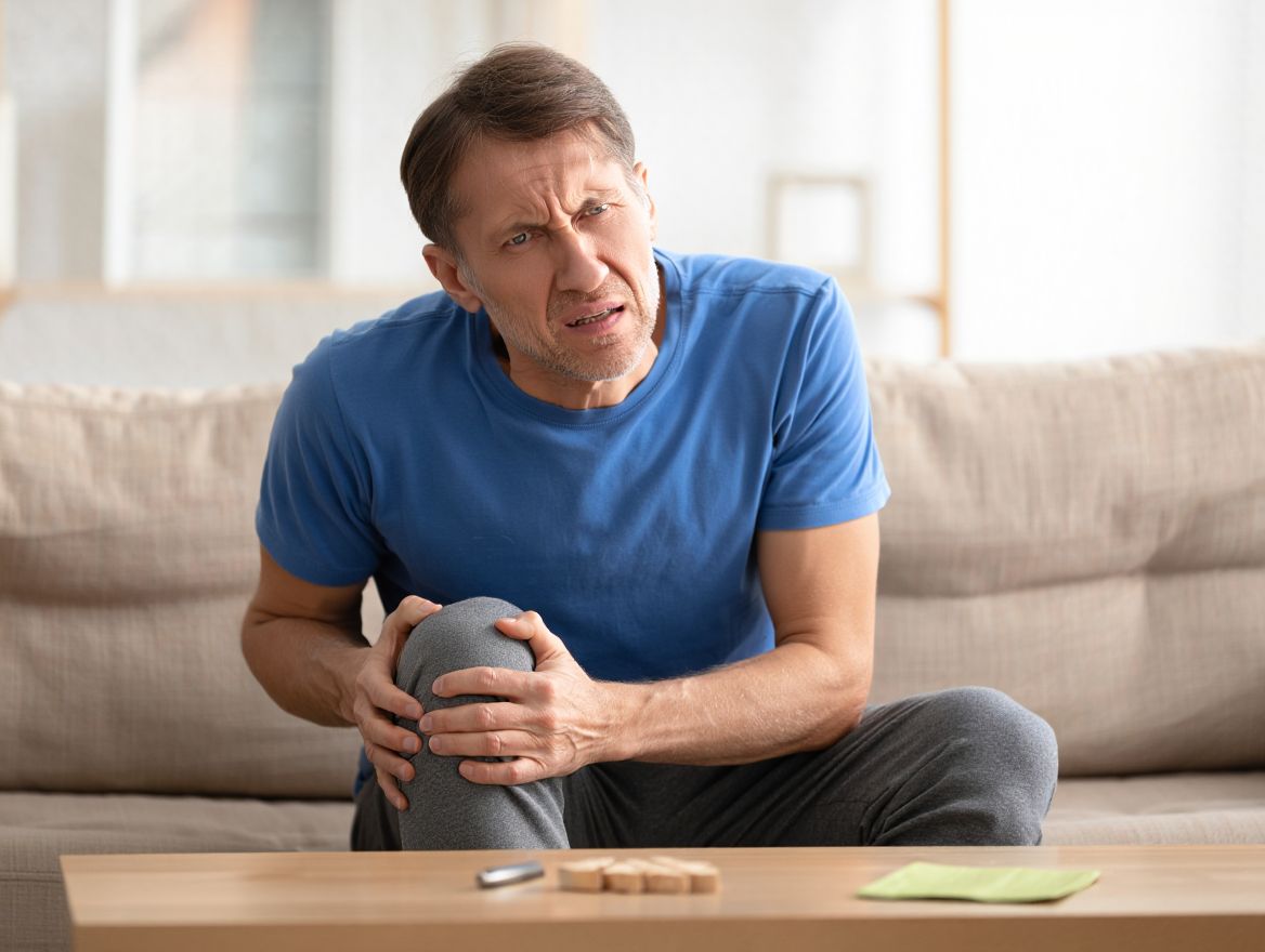 мужчина с болью в ноге сидит на диване и держится за свою ногу